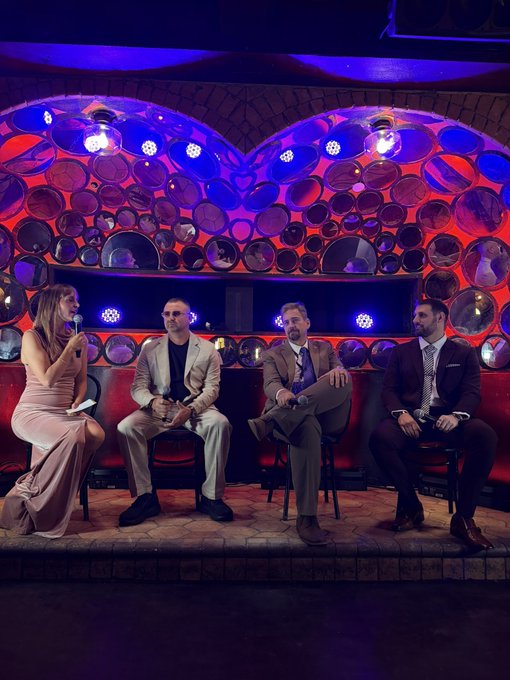 Four people sit on stools on a stage during a panel discussion, with a woman holding a microphone on the left and three men in suits beside her against a backdrop of decorative circular mirrors lit in red and blue.