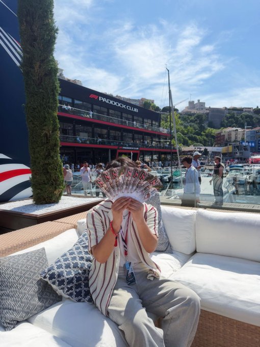 A person holds up a "MAX" branded fan in front of their face while sitting on a yacht at the Paddock Club, surrounded by the Monaco F1 harbor backdrop. The post suggests they're seeking a F1 driver's meet-and-greet invitation, with "Ric" mentioned as still having one spot left—possibly hinting at Daniel Ricciardo.