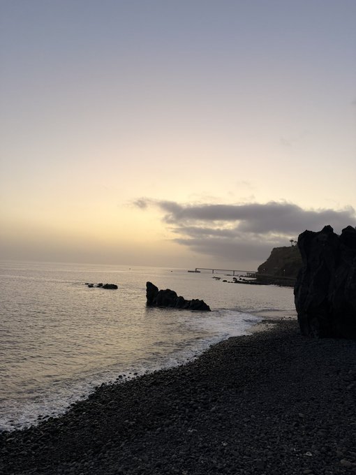 A serene sunrise over a rocky coastline bathes the calm sea and pebbled shore in soft, golden light. The tranquil scene mirrors the calm hope of a new morning, inviting viewers to share their SOL address in the dawn's quiet glow.