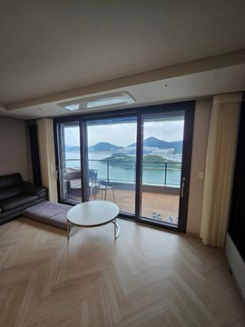 A bright living room with herringbone wood floors, a black sofa, and a round table opens through large sliding glass doors to a balcony overlooking the sea and islands.