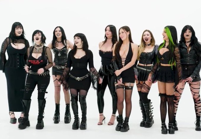A group of nine women posing together in black gothic-style outfits, including dresses, corsets, and platform boots, against a plain white background.