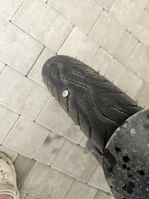 Close-up shot of a worn motorcycle tire showing damaged tread and small embedded debris, viewed from above on a tiled pavement.