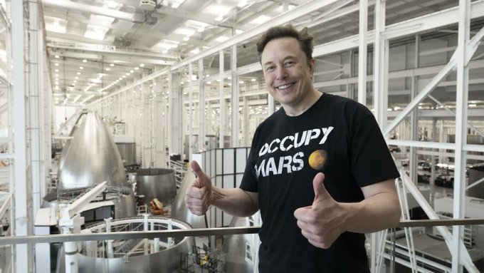 The image features a man wearing a black t-shirt with "Occupy Mars" written on it, standing inside a large building or factory. The man has his arms crossed and is giving two thumbs up, indicating that he is happy about something related to the "Occupy Mars" campaign. In the background of the photo, there are several machines and equipment, suggesting that this might be an industrial setting where the campaign's message could be promoted or implemented.