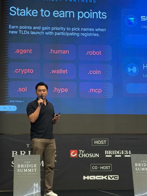 A man speaks into a microphone on stage at 'THE BRIDGE SUMMIT,' presenting a slide titled 'Stake to earn points' featuring domain extensions like '.agent,' '.human,' '.robot,' '.crypto,' and '.wallet.'