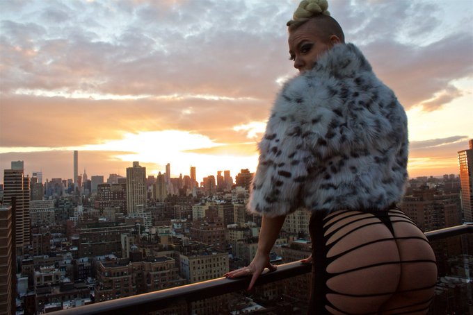 A person with a braided top-knot, wearing a faux-fur coat and cutout leggings, poses on a rooftop overlooking the New York City skyline at sunset. The image serves as the visual for the NFT titled "Top Of The World Ma."