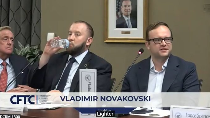 A screenshot from a CFTC (Commodity Futures Trading Commission) hearing titled 'DCRM 1300' showing Vladimir Novakovski, identified by the on-screen caption, drinking water while seated at a panel with nameplates for 'Vladimir' and 'Vance Spenc' visible.