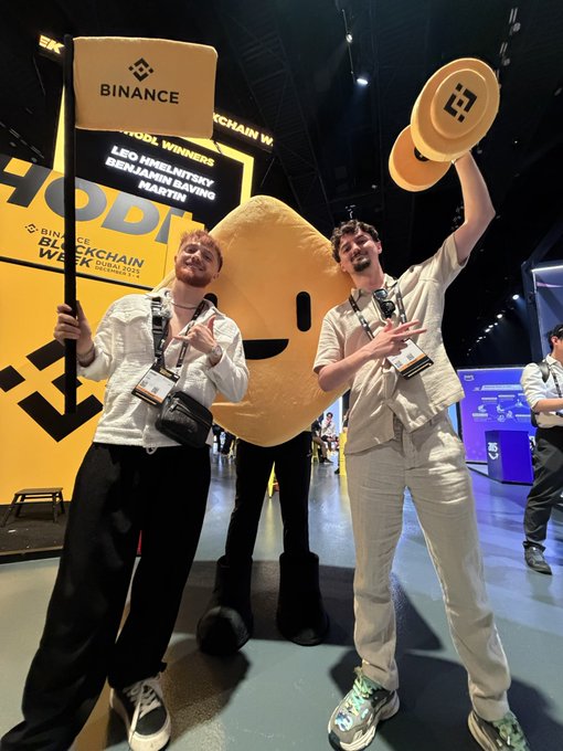 Two young men pose enthusiastically at the Binance Blockchain Week Dubai 2025 event, flanking a large Binance-branded mascot and one holding a sign displaying names of "LEO KIRILOV/SKY," "BENJAMIN BAVING," and "MARTIN" as blockchain week winners. The upbeat photo aligns perfectly with the celebratory "GM" (good morning) crypto greeting, capturing a fun, triumphant moment at the conference.