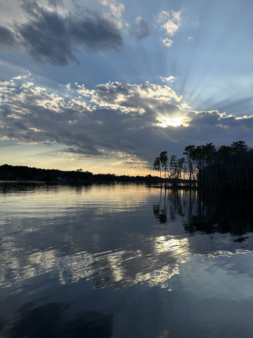 A serene landscape showing a lake reflecting a cloudy sky with sun rays beaming through, and silhouetted trees lining the shore at sunset.