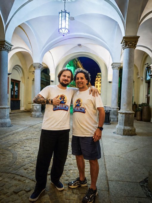 Two men stand smiling with arms around each other under a stone arcade with vaulted ceilings and columns; both wear matching white t-shirts featuring a 'CHUCK' logo with a cartoon character.