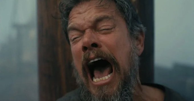 A close-up shot of a bearded man with his eyes closed and mouth wide open, appearing to scream or yell intensely.