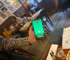 The image shows a person holding up their phone displaying a green screen with a message that reads "Just paid using Bitcoin for coffee and dessert in the historic Cafe Brasilero (1877) in Montevideo, Uruguay after visiting the Numismatico Museo at the Uruguayan Central Bank. Old versus new money!". The person is sitting at a table in a restaurant or cafe setting with chairs around them.