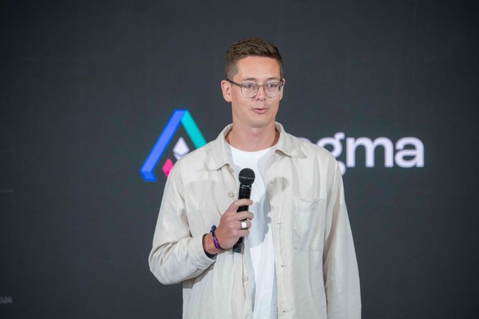 A man wearing glasses and a beige shirt holding a microphone while speaking at an event with a logo and partial text 'gma' visible in the background.
