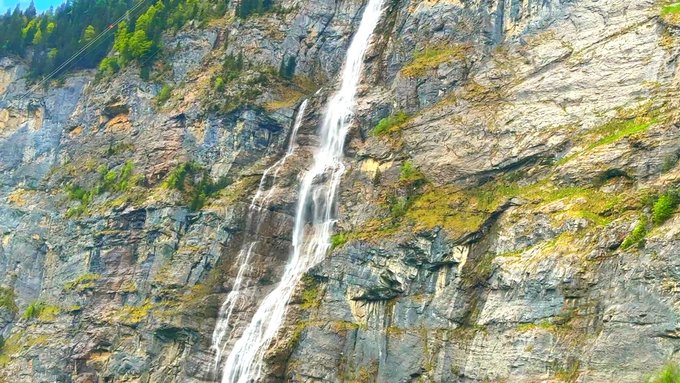 A tall, narrow waterfall cascades down the face of a rugged, rocky mountain cliff partially covered with green vegetation.
