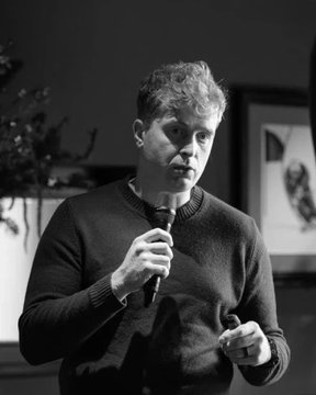 A black-and-white photograph of a man with tousled hair wearing a dark sweater, holding a microphone in one hand while gesturing with the other, appearing to give a presentation.