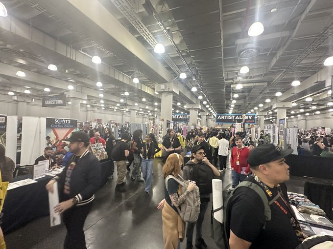 A crowded convention floor with attendees walking between vendor booths, with signs reading 'SANDSTORM', 'SCOUTS', and 'ARTIST ALLEY' hanging from the ceiling.