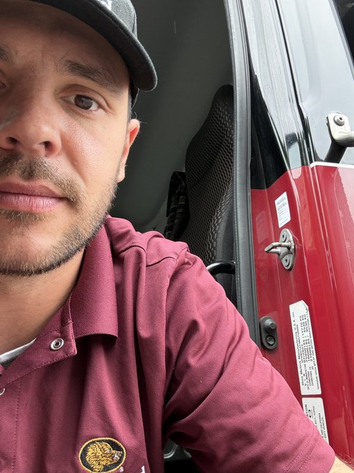 A close-up selfie of a man wearing a maroon polo shirt and a gray cap sitting inside a red vehicle, with the door and window frame visible.