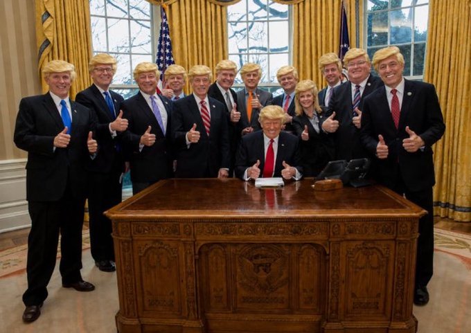 A group photo inside the Oval Office showing one person seated at the Resolute Desk while about ten others stand behind giving thumbs up, all wearing identical blonde Trump-style wigs and business attire.
