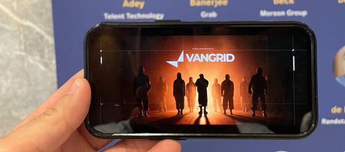 A hand holds a smartphone displaying a 'VANGRID' app screen with silhouettes of people, set against a backdrop featuring names like 'Adey' and 'Banerjee'.