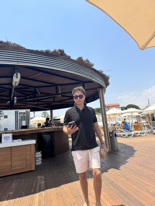A young man in a black polo shirt, pink shorts, and sunglasses stands on a wooden deck at a beach club, holding a phone and a drink.