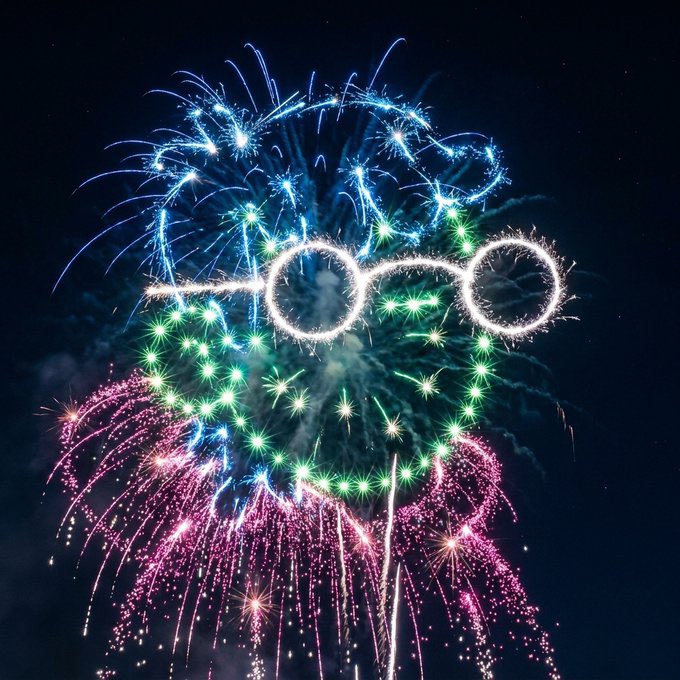 A vibrant display of fireworks in blue, green, and pink forming the shape of an Audi car logo against a dark night sky.