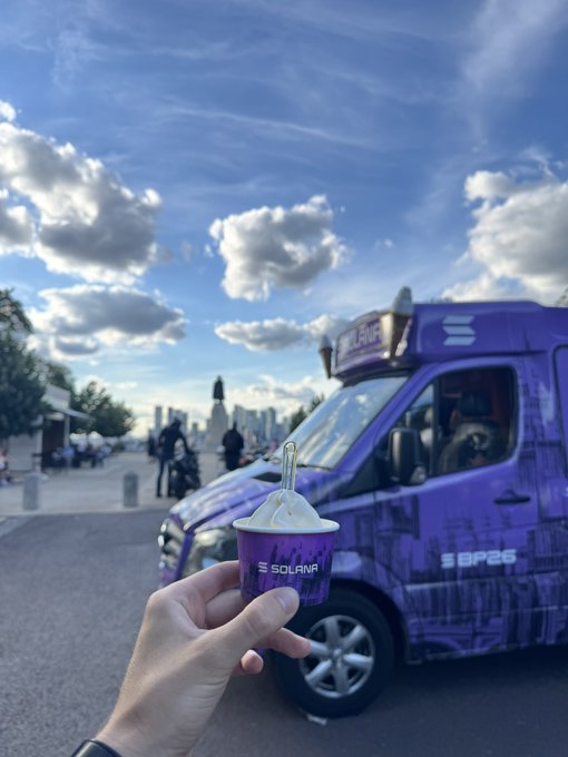 A first-person photo of a hand holding a purple 'Solana' branded ice cream cup in front of a purple 'SBP26' branded food truck, with a cloudy blue sky and a distant city skyline in the background.