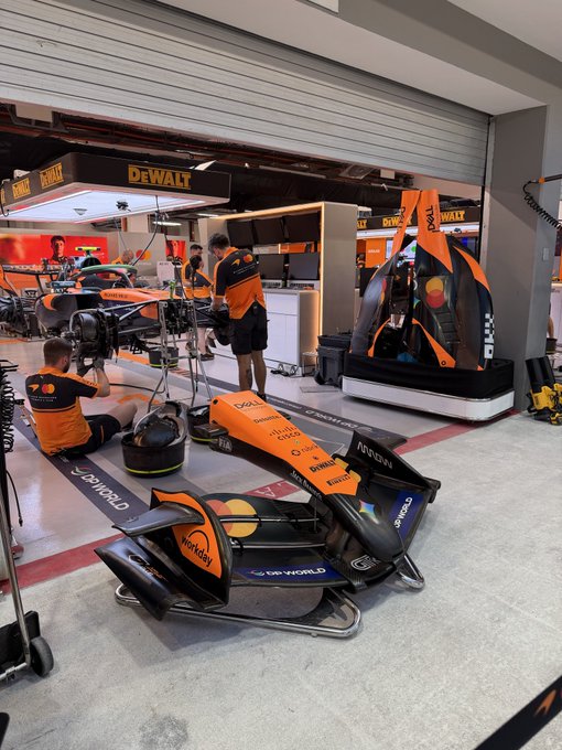 A McLaren Formula 1 team garage setup, showing a front wing assembly on the floor alongside crew members in orange and black uniforms, with DeWalt branding prominent in the background.