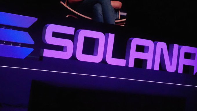 An illuminated purple 'SOLANA' sign with a logo, likely taken at a cryptocurrency or tech event, with a person sitting on a platform above it.