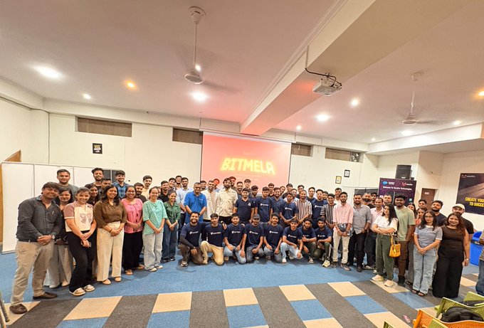 A large group of approximately 100 attendees poses together in a conference room beneath a glowing neon "#BITMELA" sign, with several participants wearing matching branded t-shirts representing the hosts of the first #RoadToBitMela event in Jaipur, sponsored by Cake Wallet.