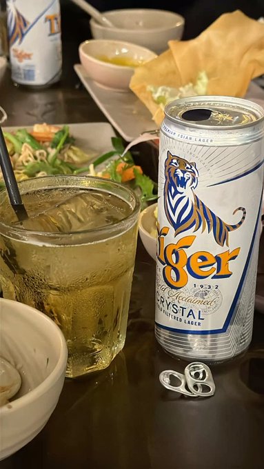 A can of 'Tiger Crystal' beer sits on a table next to a glass filled with the beverage, along with plates of food.