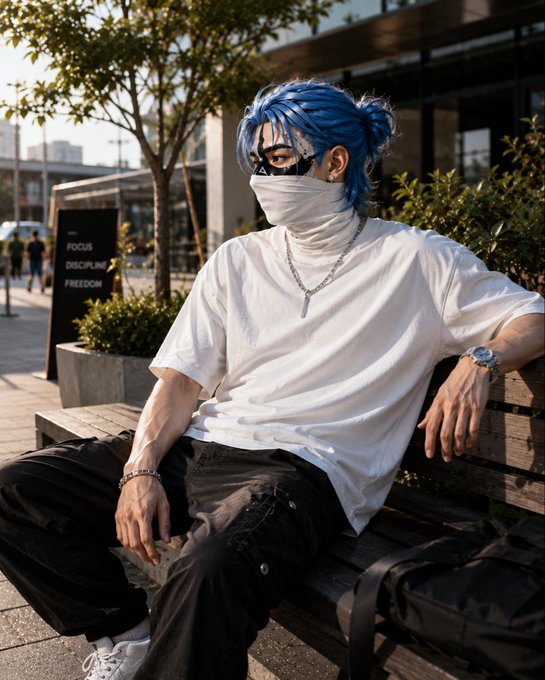 A young man with blue hair, a face mask, and a white t-shirt sits on an urban bench, accessorized with silver jewelry.