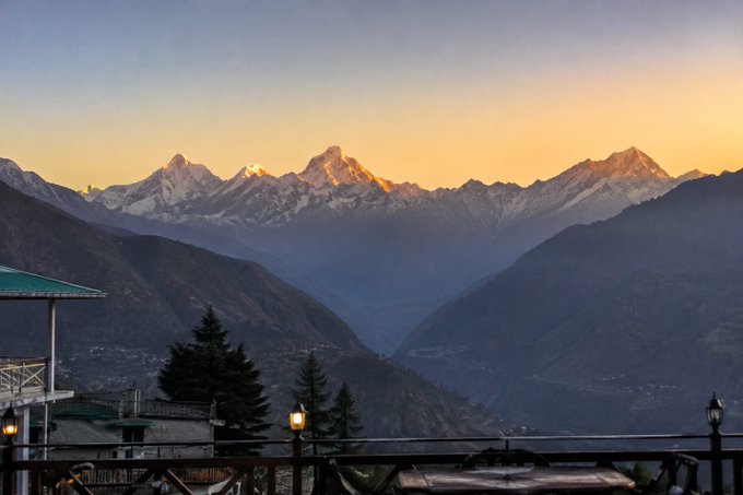 A scenic sunset view of snow-capped Himalayan mountains with a village nestled in the valley below, viewed from a balcony with a lamp post in the foreground.