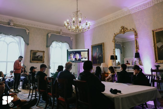 A group of people seated at tables in an ornate room watching a video call on a screen, with chandeliers, large mirrors, and classical paintings visible in the background.