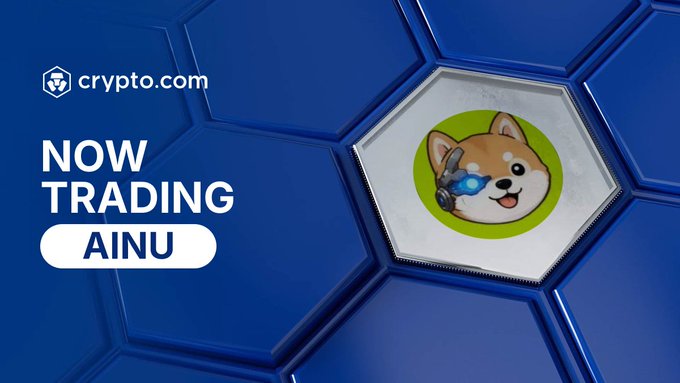 A promotional graphic from Crypto.com announcing that the token "AINU" is "NOW TRADING," featuring the project's mascot—a cartoon Shiba Inu dog wearing futuristic goggles—displayed on a blue hexagonal background alongside the Crypto.com logo.