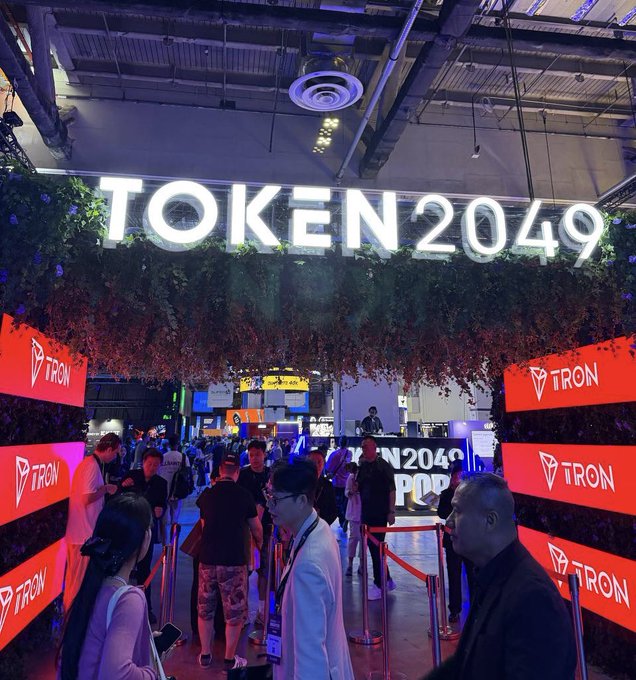 A crowded event space features a large illuminated 'TOKEN2049' sign surrounded by foliage, with red 'TRON' branded panels lining the entryway.