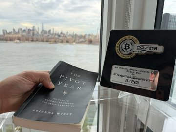 A hand holds open Brianne Wiest's "The Pivot Year" book beside a window overlooking a city skyline, with a Bitcoin-branded "Crypto Merchant" badge displaying "FRACTALENCRYPT 11/213" resting nearby. The image captures a moment of quiet reflection with Bitcoin literature, visually echoing the post's theme of finding inspiration and steady guidance from Bitcoin-focused voices during a period of daily contemplation.