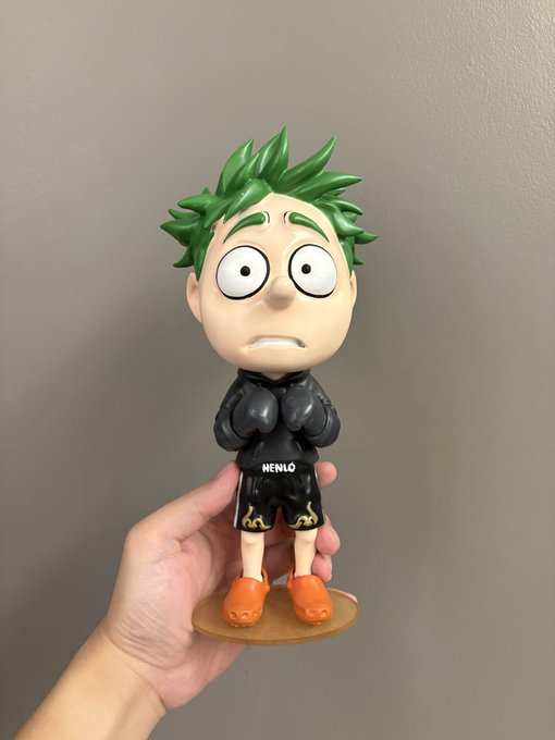 Hand-held figurine of a character with green spiky hair, large eyes, a worried expression, wearing a black outfit with 'WENLO' text and orange shoes.