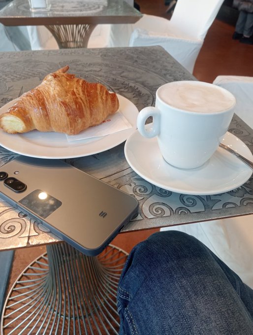 A view from a seated position showing a glass table set with a flaky croissant on a white plate, a cappuccino in a white cup, and a smartphone with a grey case. A person's legs clad in blue jeans are visible at the bottom of the frame.
