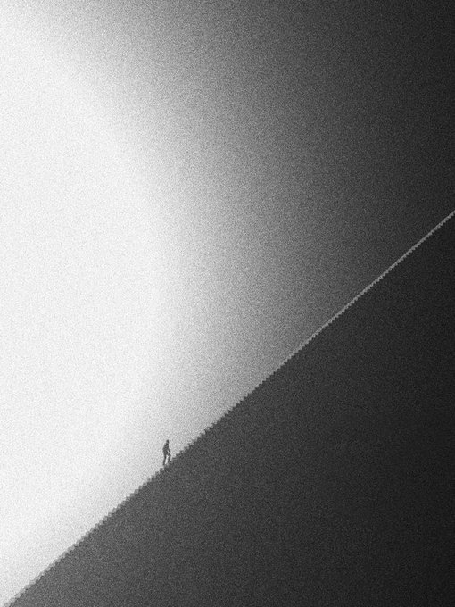 A monochrome, grainy minimalist image shows a tiny silhouetted person standing on a steep diagonal slope or ridge.