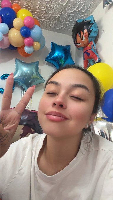 A young woman with her eyes closed and lips pressed together, making a peace sign, posing in front of a festive background of colorful balloons and a Dragon Ball-themed character balloon.