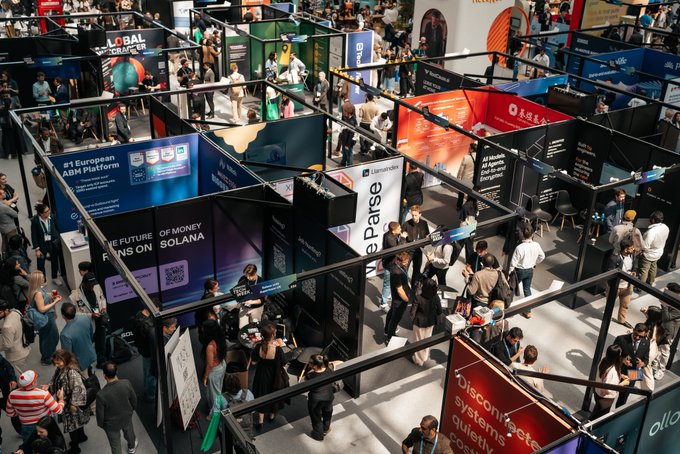 The image shows an overhead view of a crowded convention center with people walking around and exploring various booths. The main focus of the scene is a booth that has been rented out for use by a company named Solana, which seems to be attracting attention from attendees at the event. The post also mentions that Day 2 has been a major success, indicating that the conference has been well-attended and popular among participants.