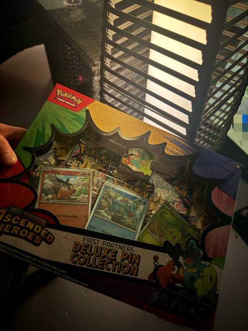 A hand holds a "First Partners Deluxe Pin Collection" Pokémon trading pin set from the "Ascended Heroes" series, featuring pins like Eevee and Marshtomp. The image ties into the post's reference to finding Pokémon merchandise "in the wild" during Pokémon Day.