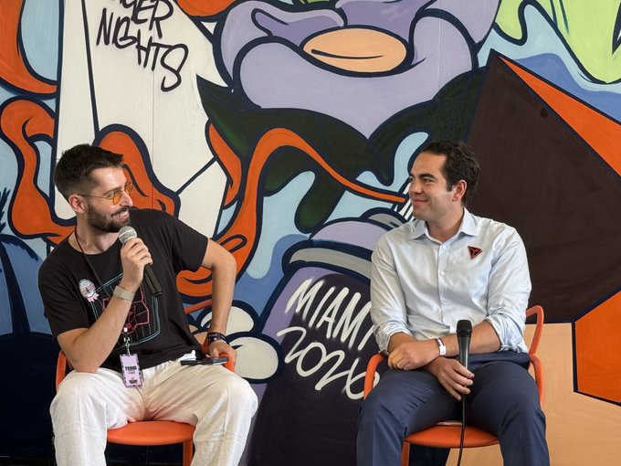 Two men sitting on orange chairs holding microphones, engaged in conversation in front of a colorful mural with 'MIAMI 2025' visible.