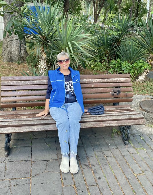 An older woman wearing sunglasses, a blue Nike vest, and jeans sits on a wooden park bench with a small purse beside her.