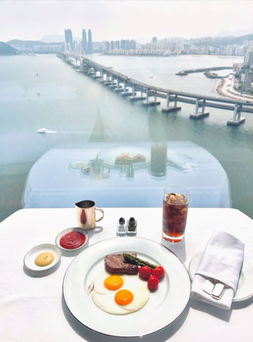 The image shows a breakfast scene with a plate of food placed on a table overlooking a body of water. The meal includes eggs, tomatoes, and ketchup. There are also two cups present - one containing tea or coffee and another filled with soda. A spoon is located near the edge of the table. The overall setting appears to be an outdoor dining area, possibly at a restaurant or a similar establishment.