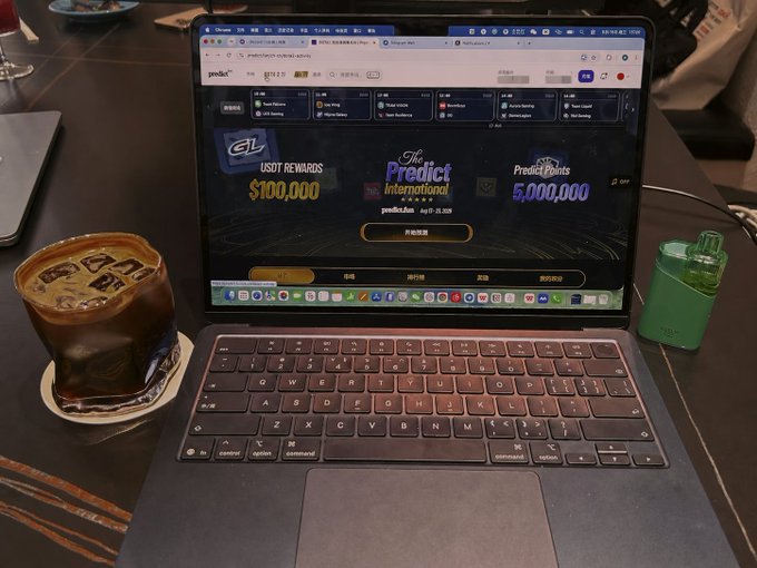 A laptop displaying a website for 'Predict International' (predict.fun) showing USDT rewards of $100,000 and Predict Points of 5,000,000, with an iced coffee and a green vape device beside it on a table.