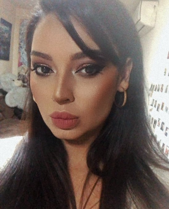 A close-up selfie of a young woman with long dark hair, bold makeup, hoop earrings, and mauve lipstick.