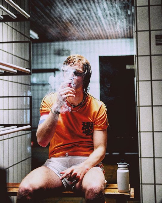 A young man in an orange Holland jersey sits in a tiled locker room smoking, likely a candid pre- or post-match moment at a Dutch football match. The "Cruyff you imbecile..." post suggests frustration with Johan Cruyff or the Dutch team, with the image capturing the gritty, raw atmosphere of a matchday.