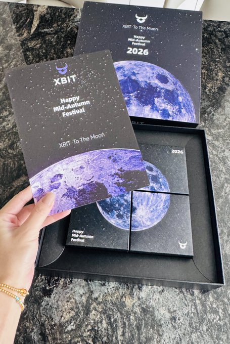 A photo of a hand holding a 'XBIT' Mid-Autumn Festival card over a gift box set featuring blue moon designs, with text reading 'XBIT - To The Moon' and 'Happy Mid-Autumn Festival 2026'.