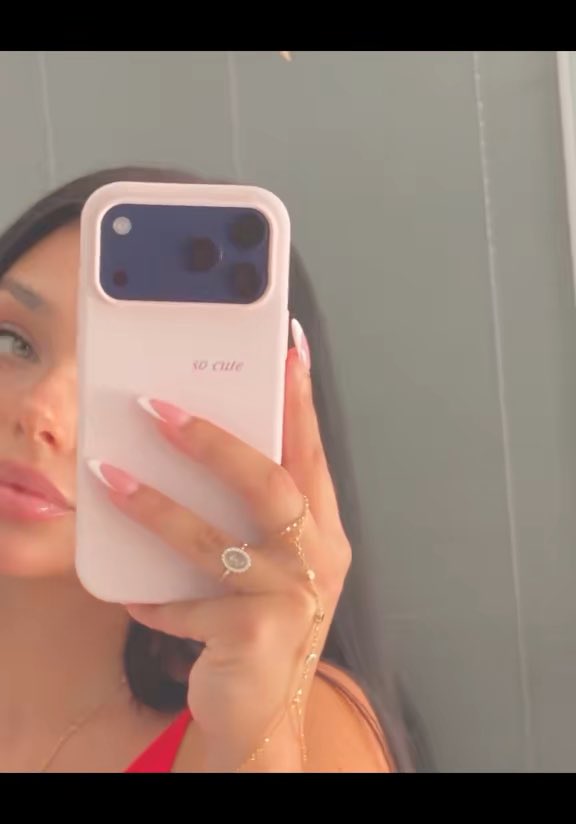 A woman takes a mirror selfie holding a pink phone case that says "so cute" on the back, wearing a delicate ring and hand chain, with the post playing on the phrase as a cheeky warning about crypto scams draining wallets after seed phrases are shared.