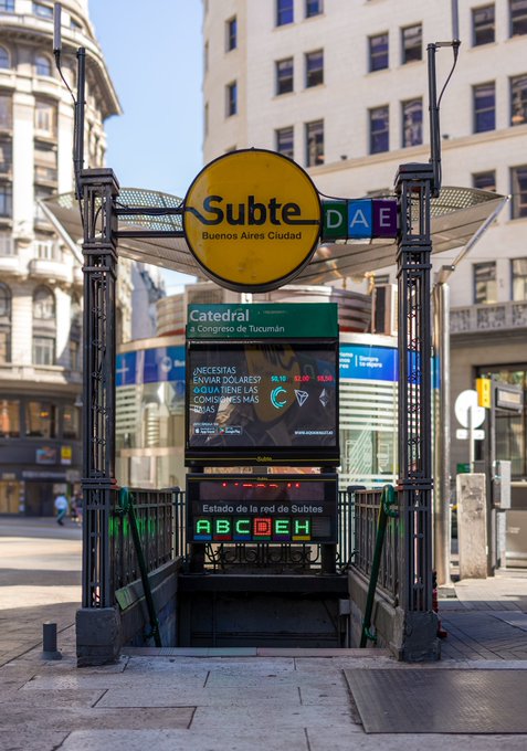 An entrance to the Buenos Aires 'Subte' (metro) at the 'Catedral' station, marked by the iconic yellow 'Subte' logo and green 'DAE' sign above the stairs leading underground, surrounded by classic city architecture.